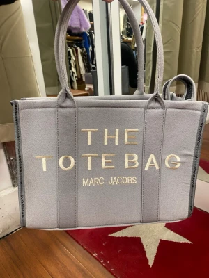 Grå shoppingväska  - Snygg shoppingväska i grå canvas . Väskan har dubbla handtag och en svart etikett med vit text på baksidan. Perfekt för dig som vill ha en trendig och rymlig tote.