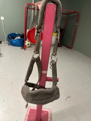 Grå grimma för ponny - Säljer en grå grimma i mjukt tyg och nylon med vadderade partier för extra komfort. Grimman har metallspännen och ringar för enkel justering och fastsättning. Märkt med 'PONY' vilket passar till ponny. Perfekt till stallet eller hagen.