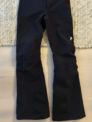 Svarta täckbyxor från Peak Performance - Säljer ett par svarta täckbyxor från Peak Performance med diskret logga på låret. Byxorna har förstärkta partier vid bensluten och är tillverkade i ett slitstarkt material som passar perfekt för vinterns alla äventyr. Snygg passform och stilren design. Lite slitna längst ner från pjäxorna 