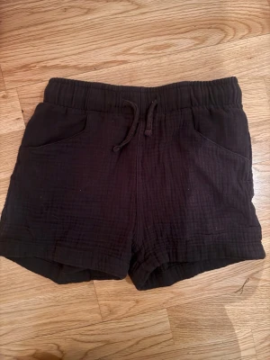 Svarta bomullsshorts med snörning - Svarta shorts i mjuk bomull med dragsko i midjan och två fickor fram. De har en avslappnad passform och är perfekta för varma dagar. Materialet är lätt och andas, vilket gör dem sköna att bära hela sommaren.