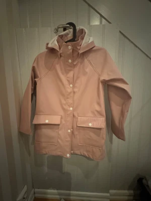 Ljusrosa regnjacka med huva - Snygg ljusrosa regnjacka med vit knäppning och huva. Jackan har två stora fickor framtill med lock och tryckknappar. Tillverkad i vattenavvisande material och har en klassisk rak passform. Perfekt för regniga dagar.