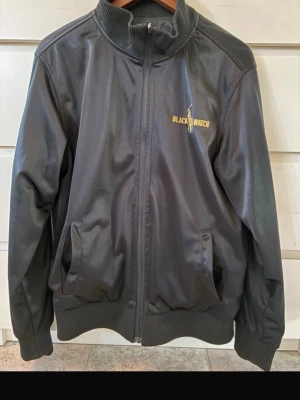 Svart vindjacka Polo Ralph Lauren - Snygg svart vindjacka från Polo Ralph Lauren med broderade detaljer i gult, bland annat texten  'Black Watch' och logga på bröstet. Jackan är vändbar och har två olika utseende. Den har ribbade muddar, hög krage och två fickor framtill. Materialet är lätt och vindtätt, perfekt för blåsiga dagar. 