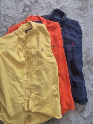 Färgglada linneskjortor från Ralph Lauren - Tre snygga linneskjortor från Ralph Lauren i färgerna gul, orange och marinblå. Alla har klassisk button-down krage, broderad logga på bröstet och slim fit passform. Perfekta för dig som vill ha en stilren och färgstark look.
