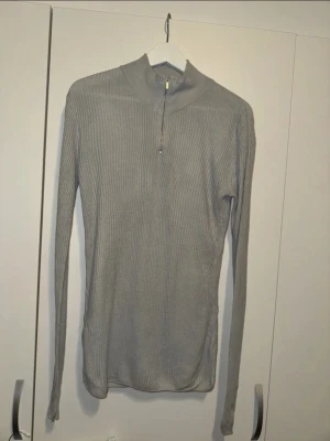 Grå ribbstickad tröja med dragkedja - Snygg grå ribbstickad tröja med lång ärm och hög krage. Tröjan har en halv dragkedja framtill och är gjord i ett mjukt, stretchigt stickat material. Enkel och clean design som funkar till det mesta.