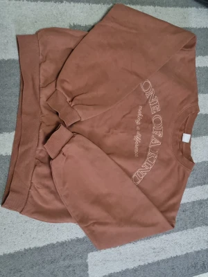 Brun sweatshirt med text - Säljer en brun sweatshirt med texten 'ONE OF A KIND making a difference' broderad på bröstet. Tröjan har rund hals, ribbade muddar vid ärmslut och nederkant samt en avslappnad passform. Perfekt för en chill och trendig look.