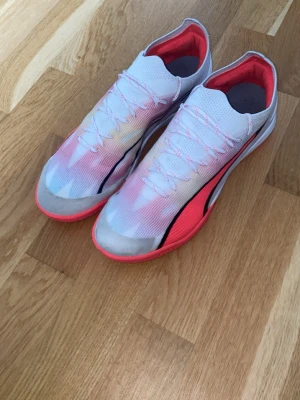 Vita Puma fotbollsskor med rosa detaljer - Säljer ett par snygga fotbollsskor från Puma i vitt med rosa och svarta detaljer. Skorna har en låg profil, tunn ovandel i syntetmaterial och snörning för bra passform. Perfekta för dig som vill ha lätta och snabba skor på planen.