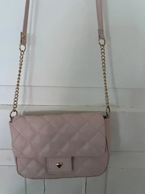 Ljusrosa quiltad axelväska med kedja - Söt ljusrosa axelväska i quiltat mönster med guldfärgad kedja och justerbar axelrem. Väskan har ett lock med tryckknapp och liten hjärtformad detalj framtill. Perfekt för dig som gillar stilrena accessoarer med en touch av romantik.