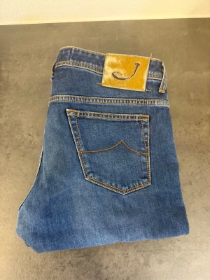 Blå jeans från Jacob Cohen, stl 35 - Säljer ett par klassiska blå jeans från Jacob Cohen med rak passform och snygga kontrastsömmar. Jeansen har fem fickor och ett gult patch med logga bak i midjan. Tillverkade i mjuk denim med en tidlös look.