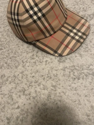 Burberry keps med klassiskt rutmönster - Snygg keps från Burberry i deras ikoniska beigea rutmönster med svarta, vita och röda linjer. Kepsen har böjd skärm och justerbar passform. Perfekt accessoar för en trendig streetstyle-look. Självklart äkta, priset kan diskuteras.