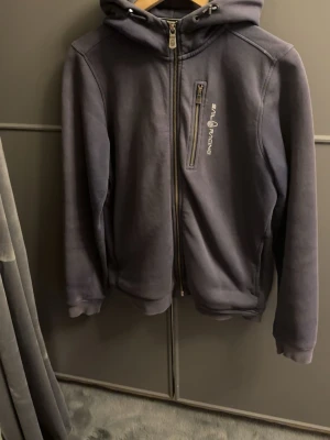 Mörkblå  hoodie från Sail Racing - Mörkgrå hoodie från Sail Racing med dragkedja framtill och bröstficka med zip. Mjuk fleece på insidan och justerbar huva med snören. Snygg logga på bröstet och sportig look. Perfekt för chill dagar eller när du vill ha en clean stil.