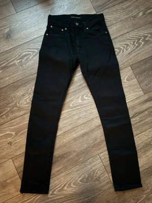 Svarta Nudie grim Tim Jeans - Säljer ett par svarta Svarta Nudie grim Tim Jeans med klassisk femficksdesign och smal passform. Jeansen har silverfärgade nitar och knapp, samt diskret logga på insidan. Perfekta för dig som gillar stilrena och tajta jeans.
