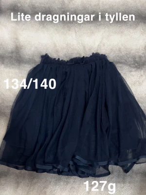 Mörkblå tyllkjol med volang - Säljer en mörkblå kjol i tyll med volangkant och resår i midjan. Kjolen har ett lager av tyll som ger ett fluffigt intryck och passar perfekt till en trendig look. 🌸 Dragningar i tyllen. ( som det blir direkt) Storlek 134/140