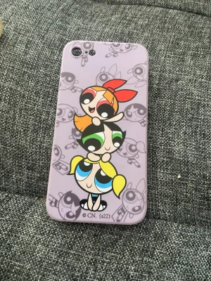 Lila Powerpuff Girls mobilskal - Säljer ett lila mobilskal med motiv av Powerpuff Girls i färgglada toner. Skalets bakgrund har ett mönster med karaktärerna i gråskala och själva trycket är i rött, grönt, blått och gult. Skal i mjukt plastmaterial som passar till iPhone 11.