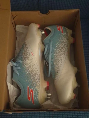 Blåvita fotbollsskor från Skechers - Säljer ett par snygga fotbollsskor från Skechers i blått och vitt med röda detaljer. Skorna har snörning, metall-dobbar och en modern design med mönstrad ovandel. Perfekta för dig som vill sticka ut på planen!