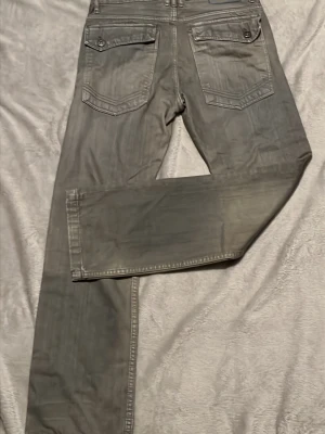 Grå jeans med stora bakfickor - Säljer ett par grå jeans med raka ben och stora bakfickor med lock och knapp. Jeansen har klassisk femficksdesign och markerade sömmar. Perfekt för dig som gillar en enkel och stilren look.