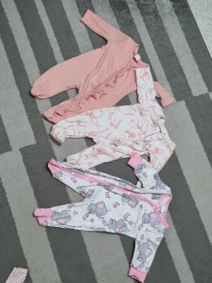 3-pack babypyjamas i rosa toner - Säljer ett set med tre babypyjamasar i mjuka material. En är enfärgat ljusrosa med volangdetalj, en är vit med rosa djur- och naturmönster, och en är vit med grå och rosa nalletryck samt rosa muddar. Alla har långa ärmar och dragkedja eller knappar framtill.