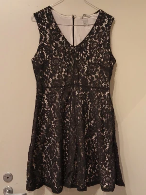 Svart spetsklänning från H&M - Säljer en svart ärmlös spetsklänning från H&M i storlek M. Klänningen har v-ringning och ett fint blommigt spetsmönster med ljus underklänning. Perfekt för dig som vill ha en elegant och stilren look.