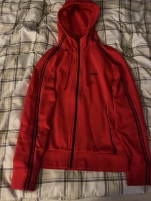 Hoodie BOSS röd - Röd hoodie från BOSS med dragkedja framtill och svarta detaljer längs ärmarna. Klassisk huva med dragsko och två fickor på sidorna. Tillverkad i mjukt material som känns skönt mot huden. Perfekt för en avslappnad och stilren look.