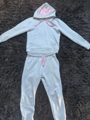 Syna World Central Cee Tracksuit - Säljer en ljusgrå hoodie med huva och rosa snören. Huvan har ett coolt rosa graffiti-tryck framtill. Hoodien har en stor magficka och är gjord i mjukt sweatshirtmaterial. Perfekt för en avslappnad och trendig look.
