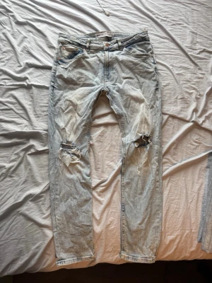 Ljusblå slitna jeans med hål - Säljer ett par ljusblå jeans med raka ben och tydliga slitningar samt hål på båda knäna. Jeansen har klassisk femficksdesign och är gjorda i denim. Perfekta för en avslappnad och trendig look.