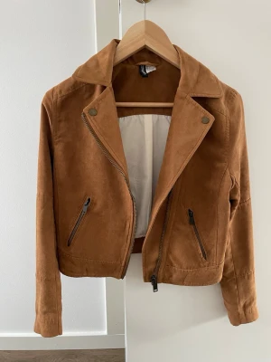 Brun jacka i mockaimitation  - Snygg brun jacka med klassisk bikerkrage och två snedställda dragkedjefickor framtill. Jackan har en kortare passform och detaljer i metall. 