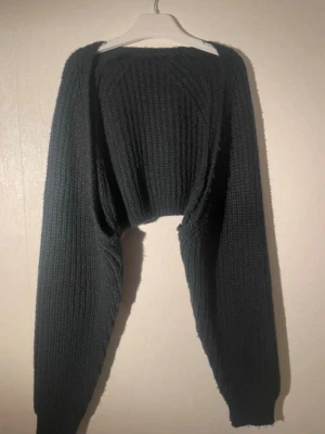 Svart ribbstickad cropped tröja - Cool svart stickad tröja med ribbad struktur och långärmad design. Tröjan är cropped och har en bred halsringning som ger en trendig look. Perfekt för att matcha med höga jeans eller kjol. Materialet är mjukt och tjockt, vilket gör den extra bekväm.