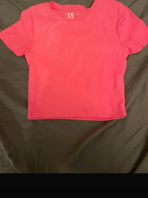 Rosa croppad ribbad topp XS - En enkel och snygg croppad topp i stark rosa färg. Toppen har korta ärmar, rund halsringning och är ribbad i materialet vilket ger en trendig känsla. Perfekt att matcha med höga jeans eller kjol.
