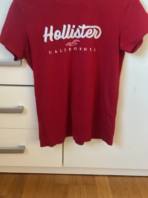Röd Hollister t-shirt med tryck - Snygg röd t-shirt från Hollister med klassiskt vitt broderat logotyp-tryck och texten 'California' på bröstet. T-shirten har rund halsringning och korta ärmar. Perfekt basic-plagg för en avslappnad stil.
