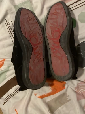 Svarta sneakers från Christian Louboutin - Snygga svarta sneakers från Christian Louboutin med den ikoniska röda sulan och diskret logga undertill. Skorna har en klassisk låg siluett och är tillverkade i svart material med matchande svarta snören. Perfekta för dig som vill sticka ut med stil.