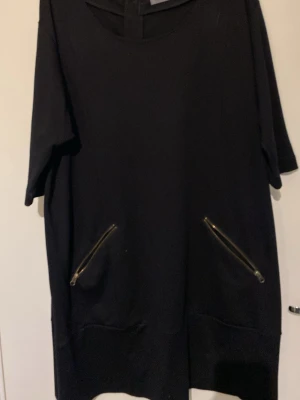 Svart kort klänning med dragkedjor - Svart kort klänning med rund halsringning och korta ärmar. Två sneda dragkedjor framtill som detaljer och en lång dragkedja bak. Enkel och stilren design, perfekt för en trendig look.