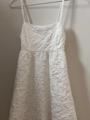 Vit kort klänning med struktur - Supersöt vit kort klänning med smala axelband och strukturerat tyg. Klänningen har en öppen rygg med knytband och en lätt utsvängd kjol. Perfekt för sommarens alla tillfällen, skolavslutning, student mm 