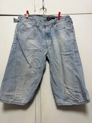 Ljusblå jeansshorts från Levi's - Säljer ett par ljusblå jeansshorts från Levi's med klassisk femficksdesign och justerbara spännen i midjan. Shortsen har en avslappnad passform och är tillverkade i slitstarkt denimtyg. Perfekta för en casual look.