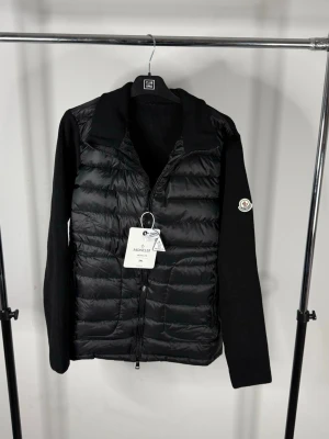 Moncler cardigan - Snygg svart dunväst från Moncler med stickade svarta ärmar och quiltad front. Västen har dragkedja framtill, hög krage och Moncler-logga på ärmen. Perfekt för lager-på-lager och ger en clean, modern look.