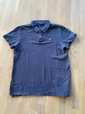 Mörkblå pikétröja från Morris - Mörkblå pikétröja från Morris med klassisk krage, två knappar framtill och broderad logga på bröstet. Tillverkad i bomull och har korta ärmar. Enkel och stilren modell. Storleken är L men sitter som M/S 