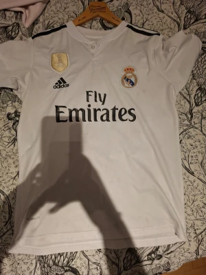 Real Madrid matchtröja Adidas vit - Vit Real Madrid matchtröja från Adidas med svarta detaljer och klubbmärke på bröstet. Tryck med Fly Emirates på framsidan och guldmärke. Klassisk sportig design med korta ärmar och rund hals. Perfekt för fotbollsträning eller att bära som supporter.
