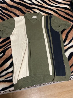 Randig pikétröja i grönt och beige - Snygg pikétröja från Urban Pioneers med breda vertikala ränder i olivgrönt, beige och mörkblått. Tröjan har krage, korta ärmar och ribbade muddar. Perfekt för en avslappnad men stilren look.