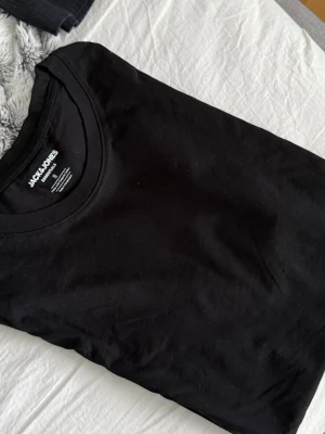 Svart basic t-shirt från Jack & Jones - En svart basic t-shirt från Jack & Jones Essentials, tillverkad i mjuk ekologisk bomull. Klassisk rund halsringning och normal passform, perfekt till alla outfits. Enkel och stilren design utan tryck eller mönster.