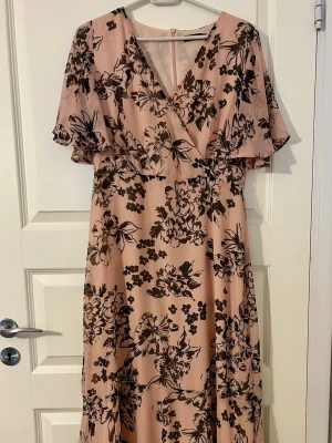 Lång omlottklänning  - Säljer en lång rosa klänning med svart blommigt mönster och omlottdesign. Klänningen har v-ringning, korta ärmar och ett flowy fall. 