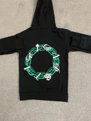 Svart hoodie Trapstar London - Svart hoodie från Trapstar London med stor grön och vit kedjegrafik på ryggen och mindre tryck på bröstet. Klassisk huva och känguruficka framtill. Snygg streetwear-stil med coola detaljer och tryck.