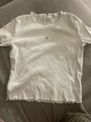 Vit ribbad t-shirt Tommy Hilfiger - Vit ribbad t-shirt från Tommy Hilfiger med liten broderad logga på bröstet. T-shirten har korta ärmar, rundad halsringning och vågig kant nertill och vid ärmslut. Enkel och stilren modell som är lätt att matcha.