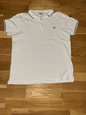 Vit piké från Gant med blå detaljer - Klassisk vit pikétröja från Gant med blå ränder på kragen och ärmsluten. Tröjan har korta ärmar, liten logga broderad på bröstet och är gjord i mjuk bomull. Perfekt till en clean och stilren look.