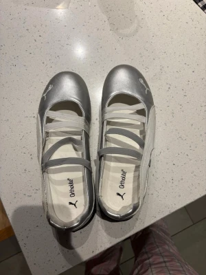 Silvriga ballerinaskor från Puma 🩶 - Snygga ballerinaskor från Puma i metallic-silver med vita detaljer och elastiska band över foten. Skorna har rund tå och Puma-logga framtill. Perfekta för dig som vill ha en sportig men stilren look. Använda kanske 5 gånger