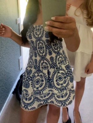 Vit och blå axelbandslös kortklänning - Axelbandslös kortklänning i vitt med blått broderat blommönster. Klänningen har en smockad överdel och volangig kjol, vilket ger en söt och somrig look. Perfekt för varma dagar och semester. Materialet känns lätt och luftigt.