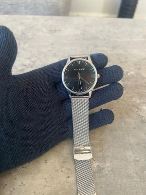 Silverklocka från Avery Milton - Stilren klocka från Avery Milton med svart urtavla och silverfärgat meshband. Klockan har en minimalistisk design med tydliga markeringar och ett klassiskt rund format. Perfekt för dig som gillar enkel och modern stil.