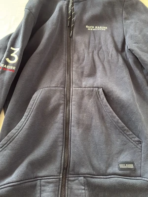 Mörkblå hoodie från Race Marine - Mörkblå hoodie med dragkedja från Race Marine. Har broderad logga på bröstet och ärmen, samt fickor framtill. Klassisk huva och ribbade muddar. Perfekt för en avslappnad och sportig stil.