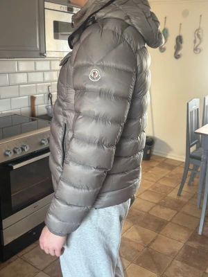 Grå Moncler dunjacka med huva - Säljer en grå Moncler dunjacka med huva och dragkedja. Jackan har två bröstfickor med knappar, broderad Moncler-logga på ärmen och quiltad design. Perfekt för kalla dagar och riktigt snygg till streetwear-stilen.