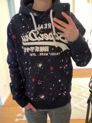 Mörkblå vintage superdry hoodie - Riktigt fin vintage superdry hoodie i mörkblå färg med röda vita och blå färgstänk på. Tröjan är storlek m och är i riktigt bra skick. Riktigt fin tröja sammanlagt. Den är lite för stor för mig men om man är längre blir passformen mer slimmad