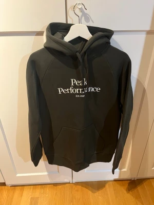Mörk grön Peak Performance hoodie - Snygg mörk grön hoodie från Peak Performance med vit logga på bröstet och klassisk känguruficka. Tröjan har huva med dragsko och är tillverkad i mjukt material som känns skönt mot huden. Aldrig använd original pris 1300kr