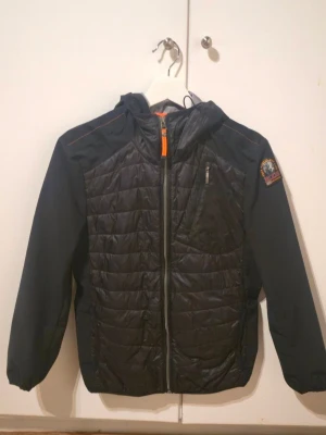 Svart vindjacka Parajumpers XS - Snygg svart vindjacka från Parajumpers i storlek XS. Jackan har quiltad front, huva, dragkedja och en bröstficka med zip. Märkespatch på ärmen. Perfekt för dig som vill ha en lätt och stilren jacka.