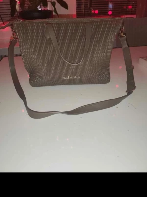 Brun handväska från Valentino - Snygg brun handväska från Valentino med flätad struktur och guldfärgade detaljer. Väskan har både handtag och justerbar axelrem samt dragkedja upptill. Invändigt finns ett fack med dragkedja och svart foder. Perfekt för dig som vill ha både stil och funktion.